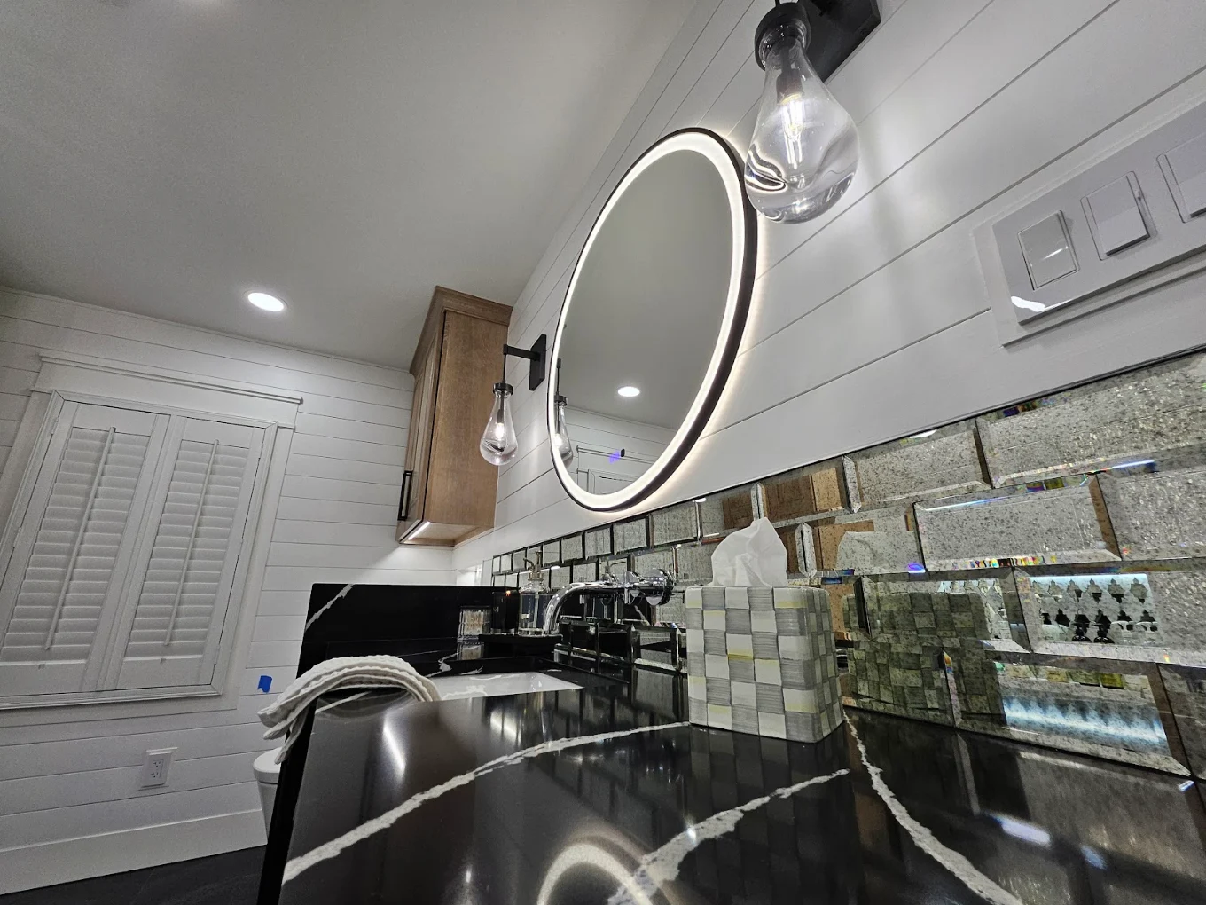 Bathroom vanity with a black countertop, chrome faucet, round illuminated mirror, and mosaic tile backsplash