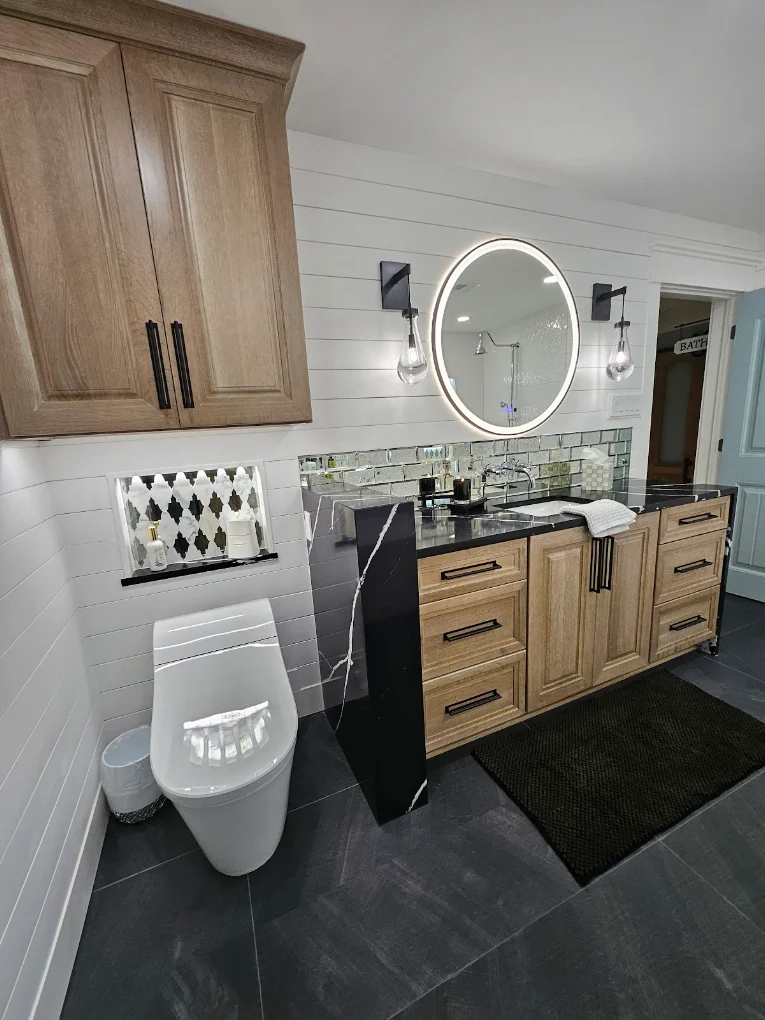 Bathroom with wood cabinetry, a black veined countertop, an illuminated oval mirror, and white shiplap walls - bathroom renovations in New Orleans